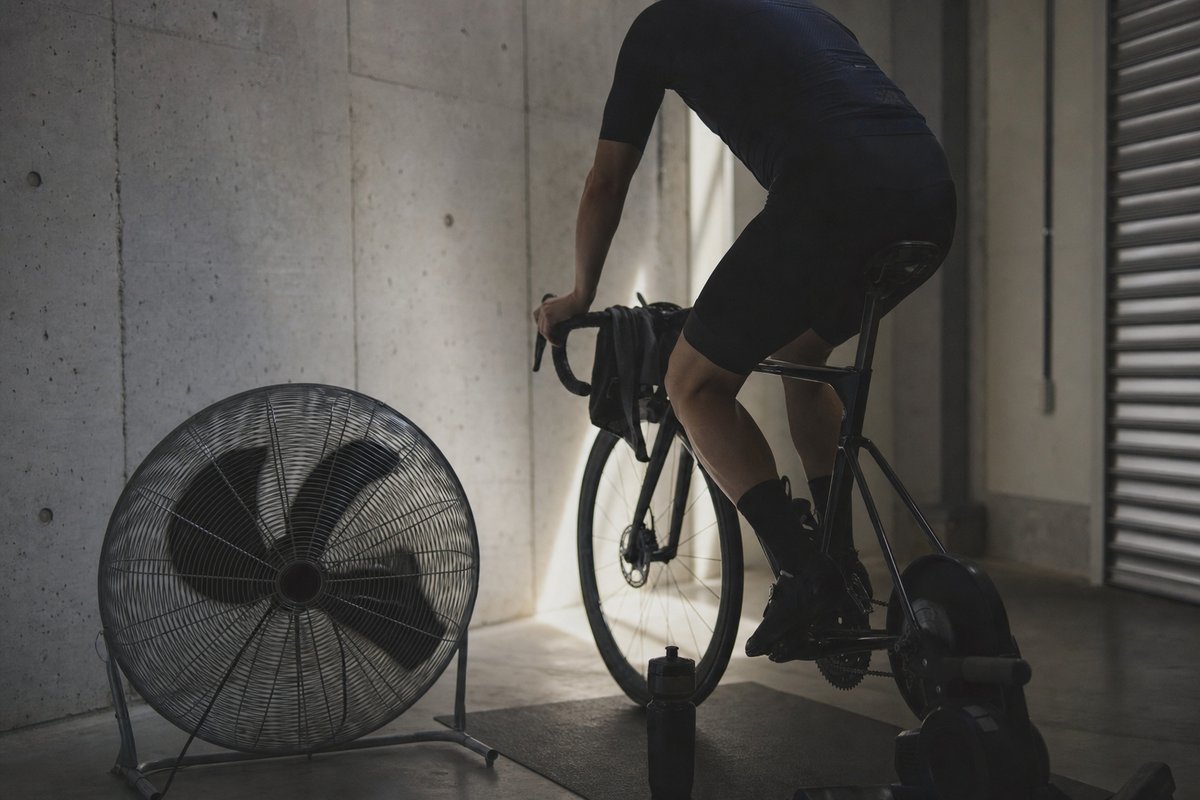 A cyclist riding hard on an indoor trainer in a bare concrete room beside a large floor fan (AI-generated image)