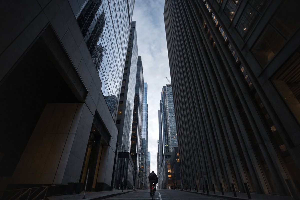 A cyclist small at the far end of a narrow street between tall glass office towers, seen from ground level (AI-generated image)