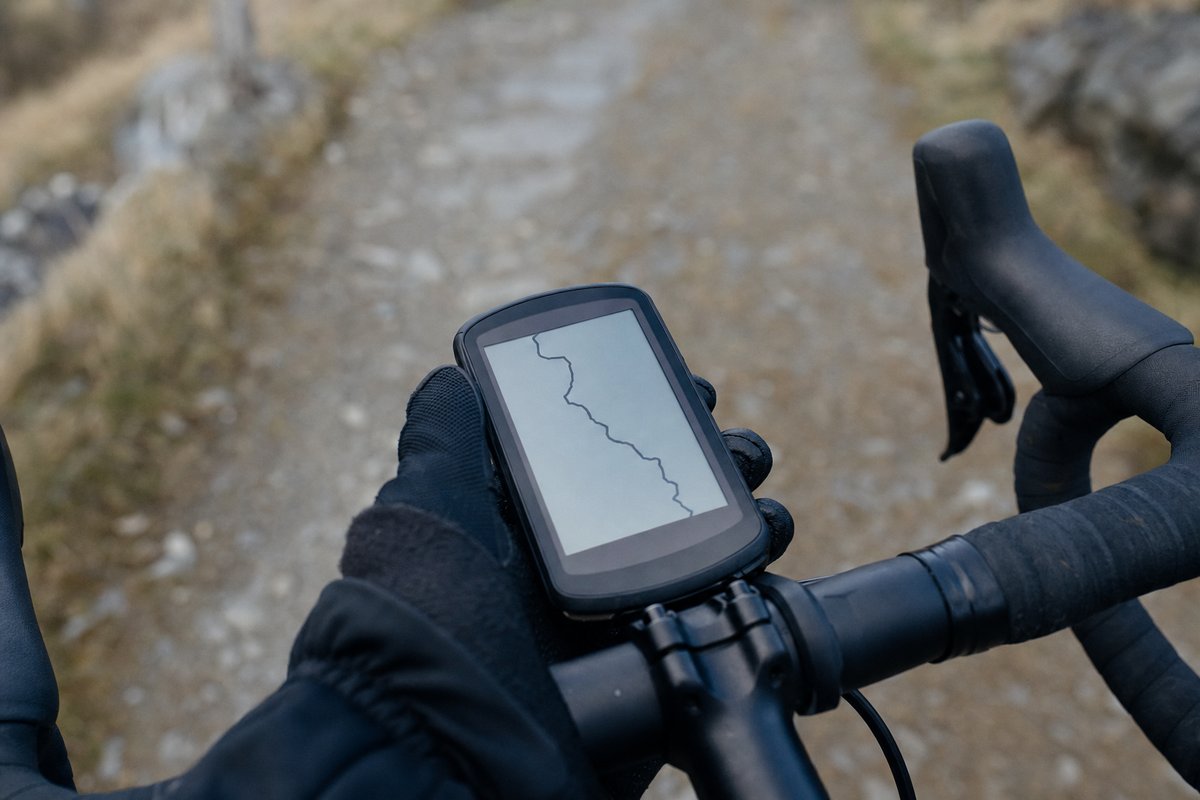 A gloved hand on drop handlebars beside a bar-mounted GPS computer showing a plain route line, a gravel track behind (AI-generated image)
