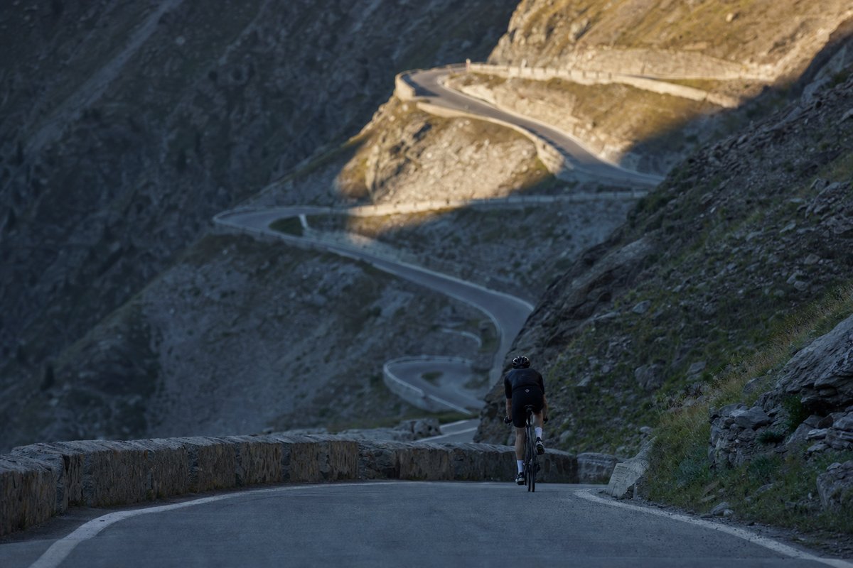 A lone road cyclist climbing a hairpin on an alpine pass, the road switching back up the mountainside behind with first light on the upper bends (AI-generated image)