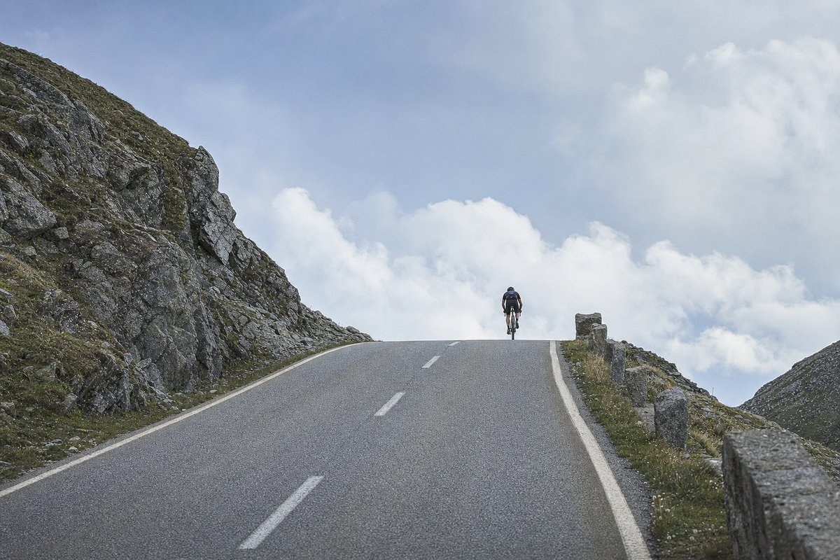 A lone cyclist cresting the top of a mountain pass road against a wide cloudy sky (AI-generated image)