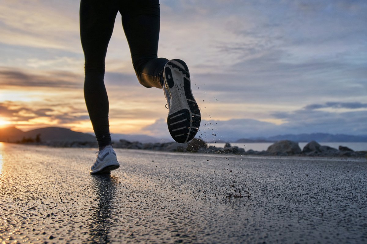 A runner's legs and shoes mid-stride on wet tarmac at sunrise, water spraying from the heel (AI-generated image)