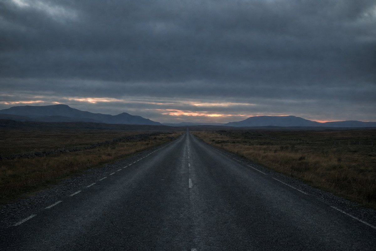 An empty road running straight across open moorland under heavy dusk cloud (AI-generated image)