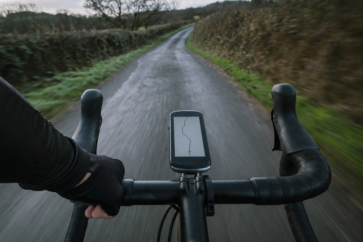 A rider's view over drop handlebars and a bar-mounted GPS computer showing a route line, the hedgerow blurred by speed (AI-generated image)