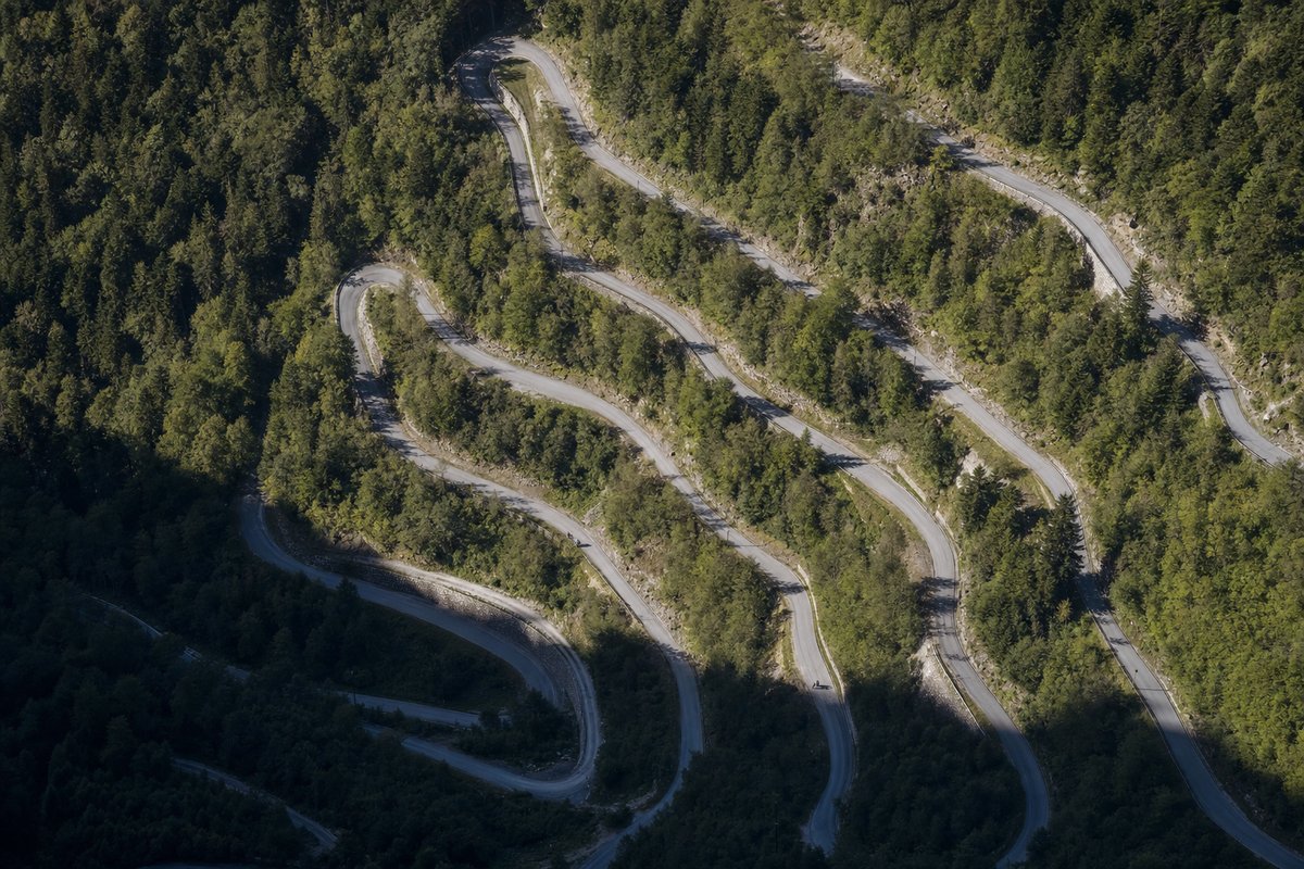 An aerial view of a mountain road switching back on itself many times up a steep forested slope (AI-generated image)